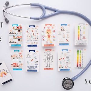 May include: A set of 10 colourful educational flashcards for kids featuring medical themes. The cards have illustrations of medical tools, body parts, and a thermometer with a temperature scale. The cards are designed to help children learn about the human body and medical care. The cards are laid out on a white surface with a stethoscope and a badge holder.