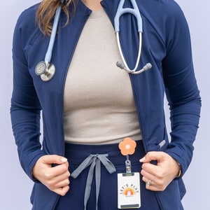May include: A woman wearing a navy blue scrub top with a light blue stethoscope around her neck. She is also wearing grey scrub trousers with a flower-shaped badge holder and a badge that says "Let's Play Badge Co."