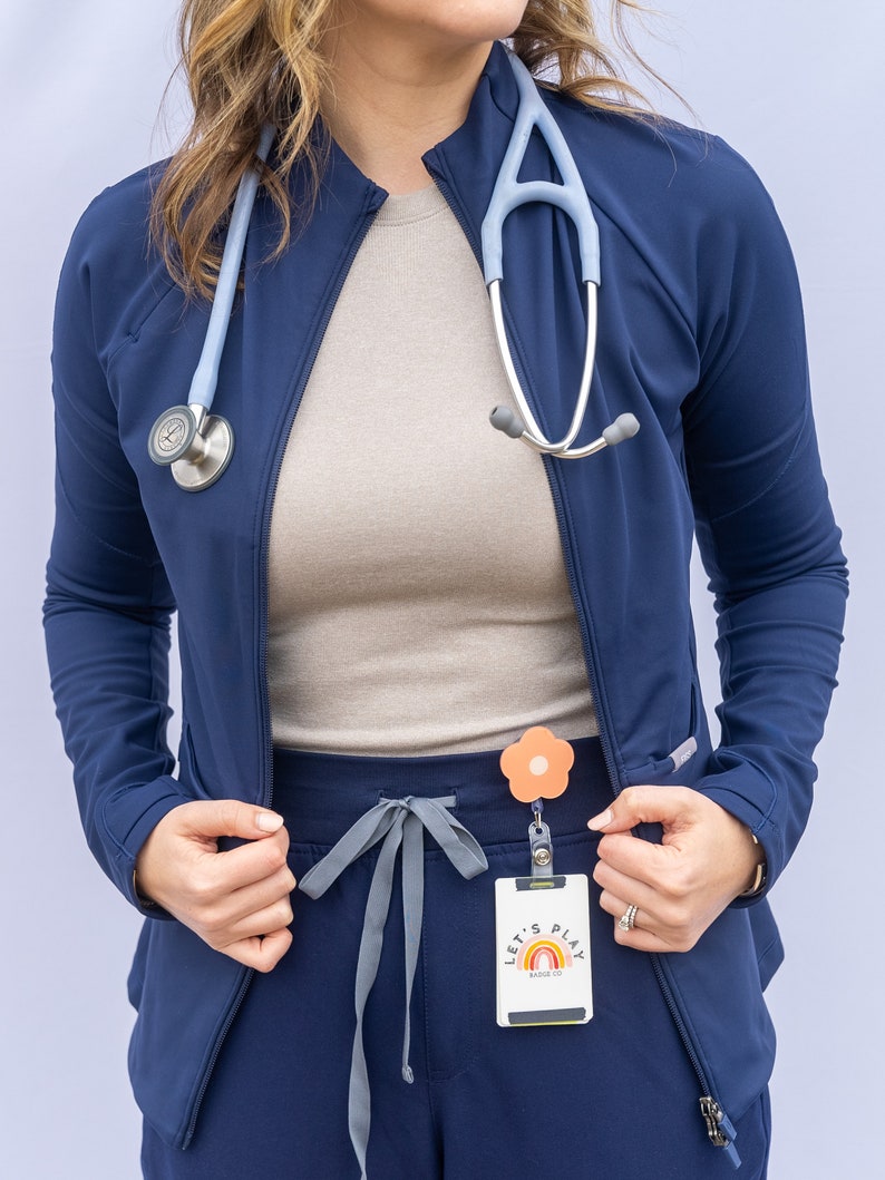 May include: A woman wearing a navy blue scrub top with a light blue stethoscope around her neck. She is also wearing grey scrub trousers with a flower-shaped badge holder and a badge that says "Let's Play Badge Co."