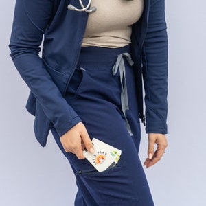 May include: A navy blue scrub top with a zip and a stethoscope hanging around the neck. The scrub top has a large pocket on the left side. A person is wearing the scrub top and holding a card that says "Let's Play" with a rainbow graphic.