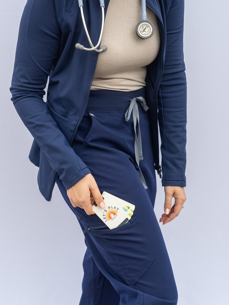 May include: A navy blue scrub top with a zip and a stethoscope hanging around the neck. The scrub top has a large pocket on the left side. A person is wearing the scrub top and holding a card that says "Let's Play" with a rainbow graphic.