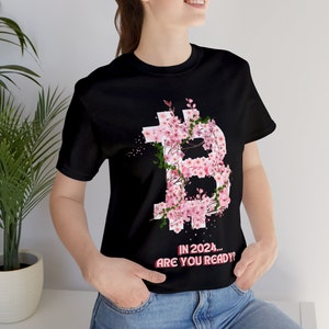 Könnte beinhalten: Schwarzes T-Shirt mit einem floralen Bitcoin-Symbol in Rosa und Weiß. Das Bitcoin-Symbol ist mit Kirschblüten verziert. Der Text "IN 2024... ARE YOU READY?" ist darunter gedruckt.