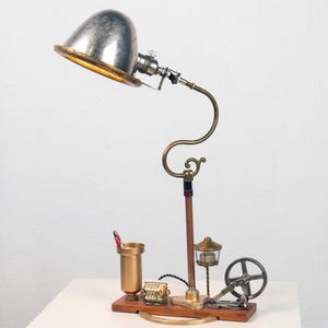 May include: A steampunk-style desk lamp with a silver dome shade, brass accents, and a wooden base. The lamp has a unique design with a curved arm and a small gear mechanism.