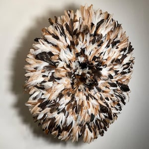 Op de afbeelding: Een ronde wanddecoratie gemaakt van veren in bruin, wit en zwart. De veren zijn gerangschikt in een radiaal patroon, waardoor een textuurrijk, decoratief stuk ontstaat. De wanddecoratie is neutraal van kleur en zou elke kamer aanvullen.