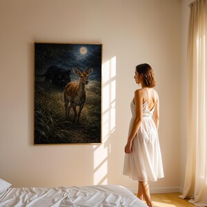 May include: A framed art print depicts a deer in a moonlit forest with a black panther in the background. The artwork is displayed on a beige wall in a bedroom setting. A woman in a white dress stands near the artwork.