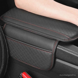 May include: Black leather car armrest cover with red stitching. The cover has a quilted pattern and is designed to provide extra comfort and support.