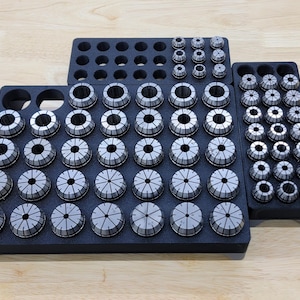 May include: A collection of silver and black collet chucks arranged in black trays. The chucks are of varying sizes and are designed for holding tools in a machining or manufacturing setting. The trays are organized for easy access and storage.