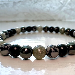 May include: A beaded bracelet featuring a repeating pattern of smooth, round beads in alternating colors of glossy black, mottled grey and white, and translucent pale green. The beads are strung together in a circular loop to form a simple wrist accessory.