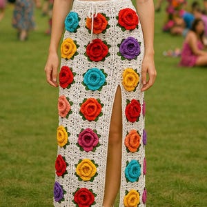 May include: A cream-colored crochet skirt with a high slit, adorned with colorful, three-dimensional rose appliqués. The roses are in shades of red, yellow, orange, pink, purple, and teal. The skirt has a drawstring waist and is worn with white sandals.
