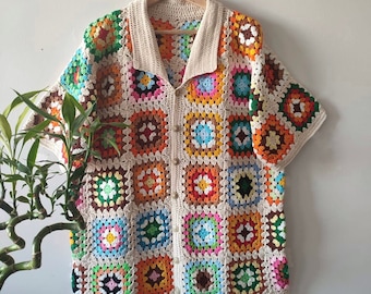 Può includere: Una camicia all'uncinetto con un motivo a quadrati della nonna colorati in tonalità di arancione, verde, blu, marrone, rosa e bianco. La camicia ha una chiusura con bottoni sul davanti e maniche corte.