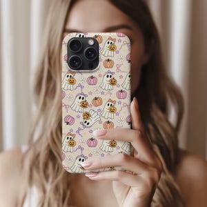 May include: A phone case featuring a repeating pattern of cartoon ghosts holding pumpkins, with pink bows, flowers, and stars on a beige background. The case has a camera cutout and is held by a person.