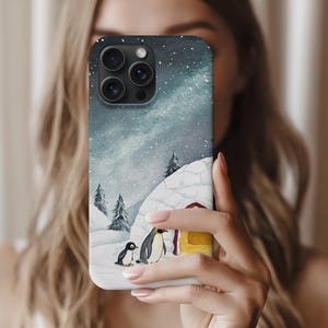 Puede incluir: Funda de teléfono con una ilustración en acuarela de un paisaje nevado con pingüinos. El diseño incluye un cielo estrellado, nieve y un pequeño iglú. La funda tiene un acabado brillante y un recorte para la cámara de lentes múltiples.