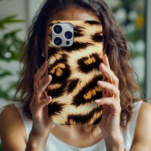 May include: A smartphone case featuring a leopard print design in shades of brown, black, and cream. The case has a glossy finish and a camera cutout. The phone case is held in front of a person's face.