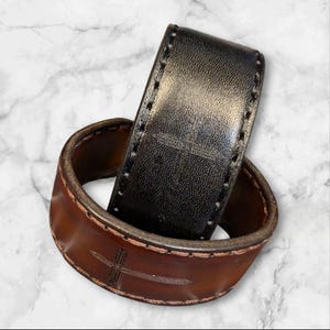 Puede incluir: Un cinturón de cuero con un diseño de cruz. El cinturón está hecho de cuero marrón y negro. El cuero marrón tiene un diseño de cruz grabado. El cuero negro también tiene un diseño de cruz. El cinturón está cosido con hilo blanco.