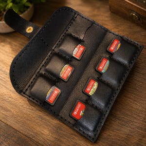 May include: A black leather game card holder featuring a fold-over flap with a gold-toned metal snap button closure. The interior contains eight individual slots, each holding a Nintendo Switch game cartridge with visible red and white labels.