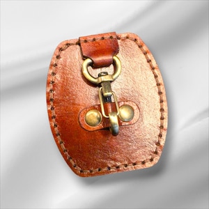 May include: A brown leather pouch with a brass clasp and fittings. The oval-shaped pouch has visible stitching around the edges. The clasp is attached to a small leather loop at the top.