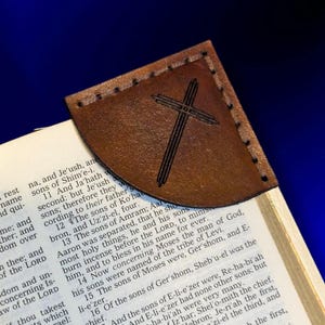 Op de afbeelding: Een bruine leren bladwijzer met een gestikte rand en een kruisontwerp. De bladwijzer is geplaatst op de pagina's van een boek, met tekst zichtbaar. De bladwijzer is een religieus item.