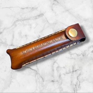 May include: A brown leather razor case with the text "SCHICK REPEATING RAZOR" embossed on the front. The case has a button closure and is stitched with a contrasting thread. The leather has a polished finish.