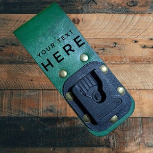 Puede incluir: Soporte de herramientas de cuero verde con el texto "YOUR TEXT HERE" en negro. Cuenta con un clip de plástico negro y está asegurado con remaches de latón. El fondo es una superficie de madera.