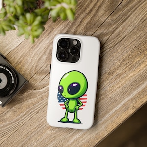 May include: White phone case with a green alien cartoon character standing in front of a red, white, and blue American flag.