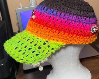 May include: A vibrant, handmade crochet baseball cap. The cap features a rainbow gradient of colors, transitioning from neon green to orange, pink, purple, and brown. Embellished with pearl accents and decorative pins, it's a unique fashion accessory.