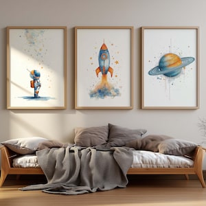 Könnte beinhalten: Drei gerahmte Aquarelldrucke mit Weltraumthemen. Einer zeigt einen Astronauten, ein anderer eine startende Rakete und der dritte einen Planeten mit Ringen. Die Drucke werden über einem hölzernen Tagesbett mit Kissen und einer Decke ausgestellt.