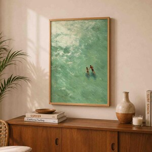 May include: A framed wall art print featuring an aerial view of two people swimming in turquoise ocean water. The artwork is displayed in a light wood frame, capturing the movement of waves and the figures of the swimmers in the center.