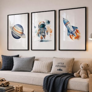 May include: Three framed wall art prints featuring watercolour illustrations of a blue planet with rings, an astronaut in a white suit, and a blue rocket ship with orange flames, all set against a clean white background in black frames.