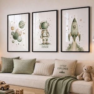 May include: Three framed wall art prints featuring watercolor illustrations of space themes. The left print shows planets, the center print depicts an astronaut with the name Oliver, and the right print displays a rocket ship with green accents.