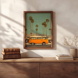 May include: A framed print featuring an orange vintage van driving past palm trees under a teal sky. The artwork is displayed on a wooden cabinet with books and a vase of dried flowers. The frame is a dark brown color.