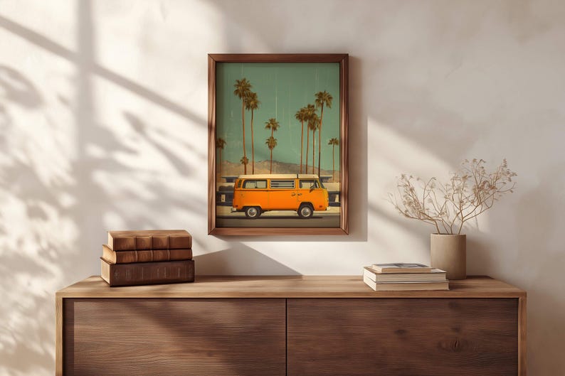 May include: A framed print featuring an orange vintage van driving past palm trees under a teal sky. The artwork is displayed on a wooden cabinet with books and a vase of dried flowers. The frame is a dark brown color.
