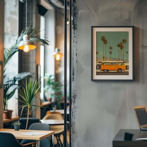 May include: A framed print featuring an orange vintage van parked on a road, with palm trees in the background under a teal sky. The artwork is displayed on a gray wall in a cafe setting, with a table and laptop visible.