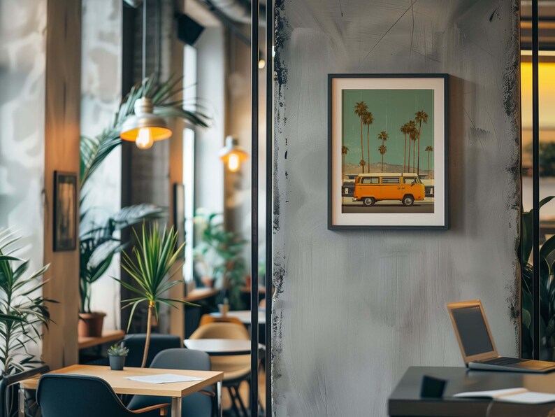 May include: A framed print featuring an orange vintage van parked on a road, with palm trees in the background under a teal sky. The artwork is displayed on a gray wall in a cafe setting, with a table and laptop visible.