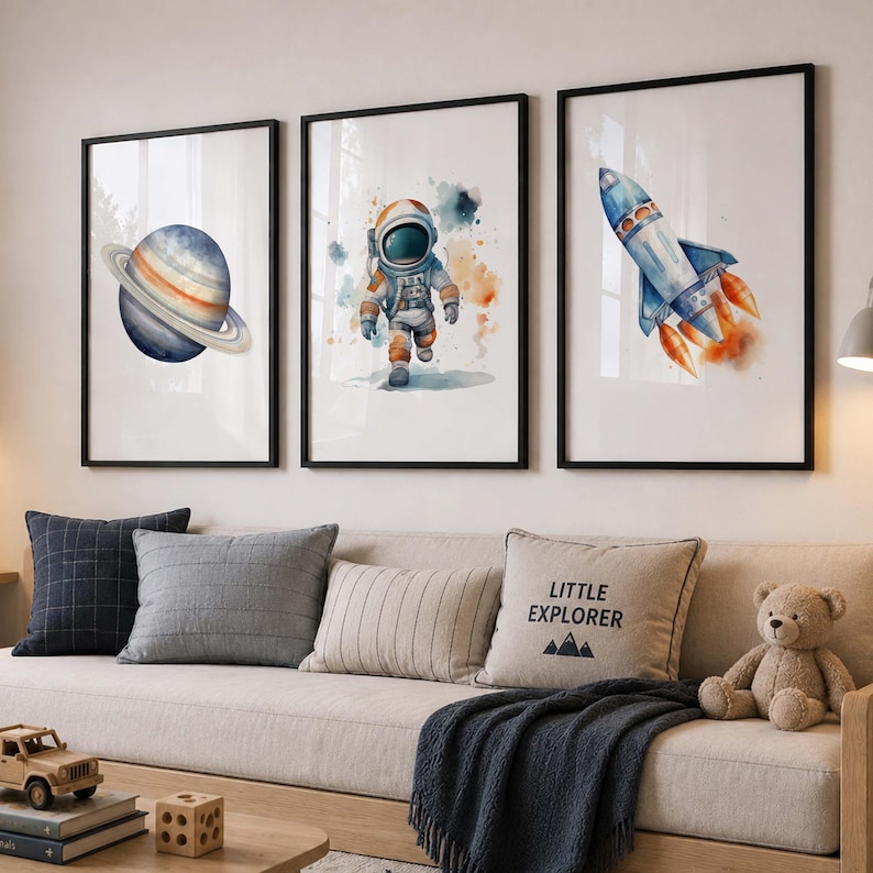 May include: Three framed wall art prints featuring watercolor illustrations of a blue planet with rings, an astronaut in a white suit, and a blue rocket ship with orange flames, all set against a clean white background in black frames.