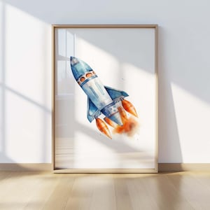 May include: A framed wall art print featuring a watercolor illustration of a blue and white rocket ship angled upward. The rocket has orange exhaust flames emerging from its base, creating a soft, blended effect against the white background.
