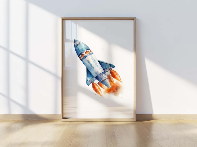 May include: A framed wall art print featuring a watercolor illustration of a blue and white rocket ship angled upward. The rocket has orange exhaust flames emerging from its base, creating a soft, blended effect against the white background.