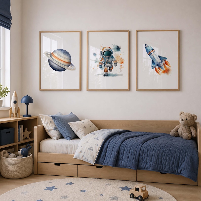 May include: Three framed wall art prints featuring watercolor illustrations of a planet with rings, an astronaut in a space suit, and a rocket ship launching with orange flames, displayed above a wooden daybed with blue bedding and a teddy bear.