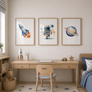 May include: Three framed wall art prints featuring watercolor illustrations of a space rocket, an astronaut, and a planet with rings. The artwork is displayed in light wood frames, showcasing vibrant blue, orange, and grey tones on white backgrounds.