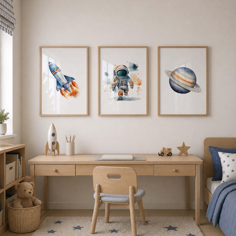 May include: Three framed wall art prints featuring watercolor illustrations of a space rocket, an astronaut, and a planet with rings. The artwork is displayed in light wood frames, showcasing vibrant blue, orange, and grey tones on white backgrounds.