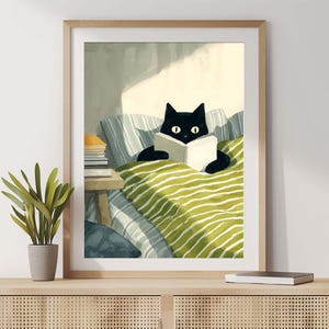 Puede incluir: Una impresión artística enmarcada que representa a un gato negro leyendo un libro en la cama. El gato está sobre una manta a rayas verdes y blancas. El fondo incluye una pequeña mesa con libros y una ventana con luz solar.