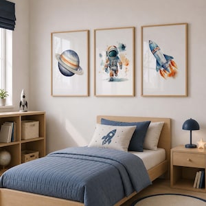May include: Three framed wall art prints featuring watercolor illustrations of a planet with rings, an astronaut in a space suit, and a rocket ship launching with orange flames, displayed in a bedroom above a bed with blue bedding.