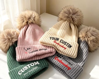May include: Four ribbed knit beanies with large faux fur pom-poms in green, pink, beige, and gray. The green beanie features white text reading CUSTOM your text-logo. The pink one has a small graphic and text reading Custom design. The beige and gray beanies feature embroidered text reading YOUR CUSTOM Embroidery.