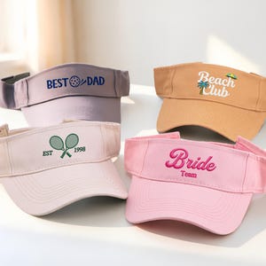 May include: Four visors in different colours are shown. One is grey with "BEST DAD" embroidered in blue. Another is tan with "Beach Club" and a palm tree. A third is pink with "Bride Team". The last is light pink with tennis rackets.