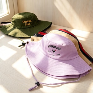 May include: A collection of bucket hats in various colors, including olive green and lavender. The olive green hat has "Custom Text" embroidered on it. The lavender hat features a cocktail glass logo with "Custom Logo Est 1998". Each hat has a chin strap.