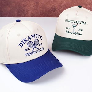 May include: Two baseball caps. One is cream-coloured with a blue brim and the text "DIKAWSYK EST. 2026 TENNIS CLUB" in blue. The other is cream-coloured with a green brim and the text "GERENARTRA EST 1998 Cherry Martini" in green.