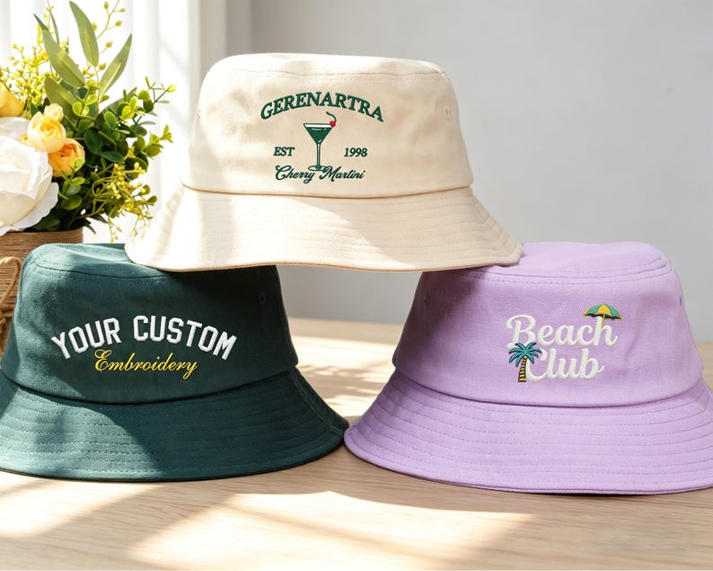 Pu&ograve; includere: Tre cappelli a secchiello color crema, verde scuro e lavanda. Il cappello color crema ha il testo "GERENARTRA" e "Cherry Martini". Il cappello verde dice "YOUR CUSTOM Embroidery". Il cappello lavanda dice "Beach Club" con una palma e un ombrellone.