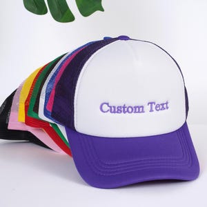 May include: A stack of colorful trucker hats with a white hat on top featuring purple text that says "Custom Text".