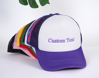 May include: A stack of colorful trucker hats with a white hat on top featuring purple text that says "Custom Text".