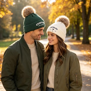 May include: A dark green ribbed knit beanie with a brown faux fur pom-pom and white text reading CUSTOM, and a cream ribbed knit beanie with a brown faux fur pom-pom and blue text reading CUSTOM LOGO with a small martini glass icon.