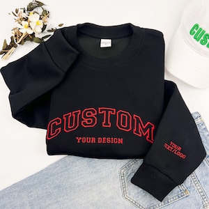 May include: A black crewneck sweatshirt featuring red embroidered text that reads CUSTOM and YOUR DESIGN on the front, with the text YOUR TEXT/LOGO embroidered in red on the sleeve cuff. The garment has long sleeves with ribbed cuffs.